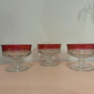 Può includere: Tre coppette da dessert vintage in vetro trasparente con bordi rosso rubino. Le coppette hanno un motivo decorativo al centro e poggiano su una base a piedistallo trasparente. Le coppette sono disposte su una superficie rosa.