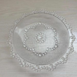 Puede incluir: Un plato de servir de vidrio transparente con una superficie texturizada y un diseño decorativo de burbujas alrededor del borde. El plato presenta un patrón central en forma de flor de burbujas. El plato es redondo y mide unos 30 cm de diámetro.