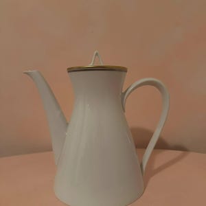 May include: White ceramic teapot with a long spout and curved handle. The lid and rim are accented with gold trim. The teapot has a classic, elegant design, suitable for serving tea or coffee.