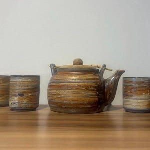 May include: A ceramic tea set with a teapot and four matching cups. The set features a brown and tan striped pattern. The teapot has a wooden handle and lid. The cups are cylindrical.