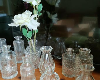 May include: A collection of ten clear glass bud vases of varying shapes and textures, including ribbed, diamond-patterned, and smooth designs. One vase contains two artificial white flowers with green stems and leaves.