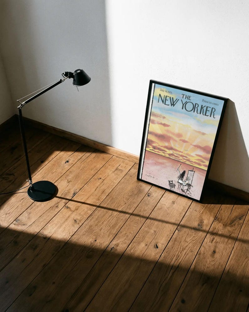 May include: A framed magazine cover of The New Yorker dated July 16, 1983, featuring a watercolor illustration of a sunset over a desert landscape with an artist painting at an easel, displayed in a black frame next to a black metal desk lamp.