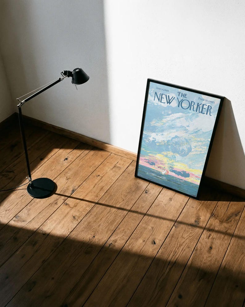 May include: A black adjustable floor lamp with a round base stands next to a framed New Yorker magazine cover. The magazine cover features a colorful sky scene with the text "The New Yorker". The items are on a wooden floor.