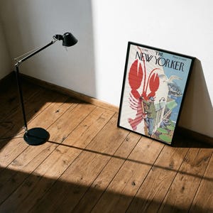May include: A framed New Yorker magazine cover with a red lobster design and the magazine's title. A black floor lamp is to the left, casting a shadow on the wooden floor. The magazine is leaning against a white wall.
