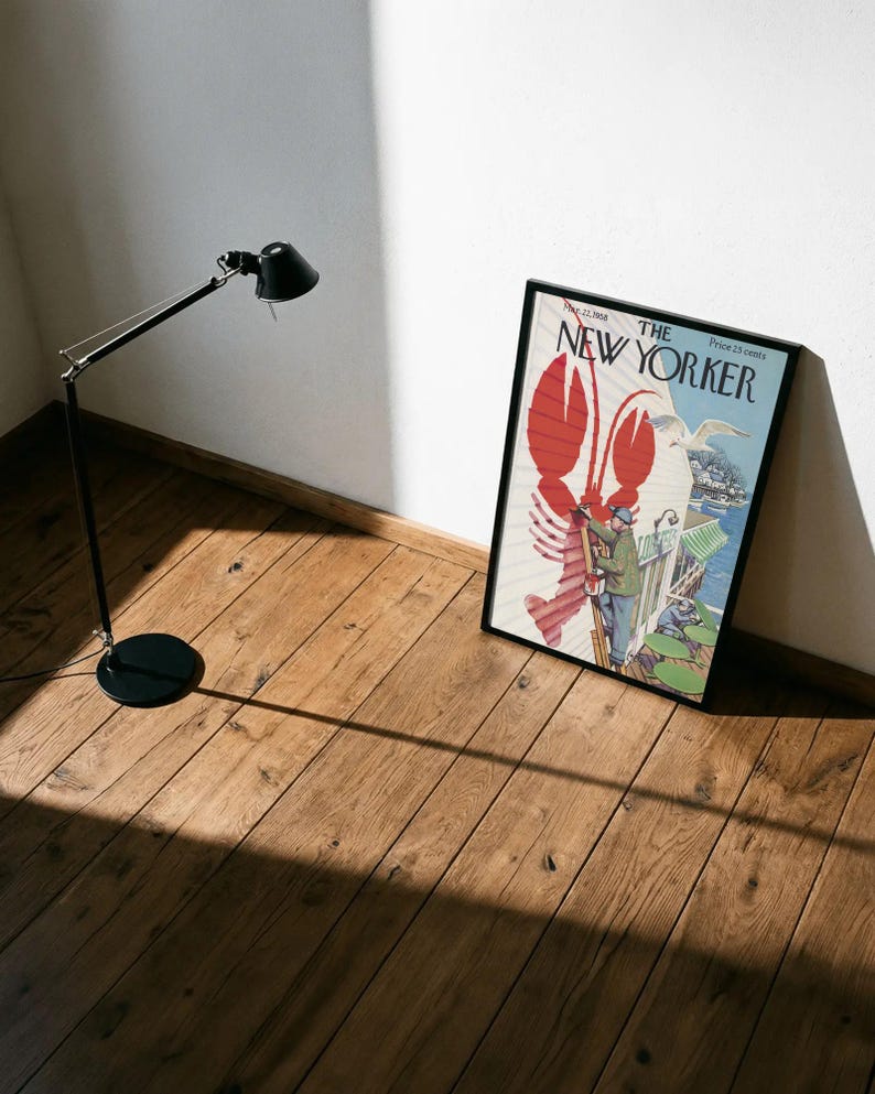 May include: A framed New Yorker magazine cover with a red lobster design and the magazine's title. A black floor lamp is to the left, casting a shadow on the wooden floor. The magazine is leaning against a white wall.