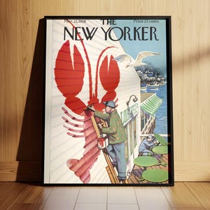 May include: Framed vintage New Yorker cover art with a large red lobster illustration. The cover art includes a man painting the lobster, a seagull, and the magazine's title. The frame is black and the artwork is leaning against a wooden wall.