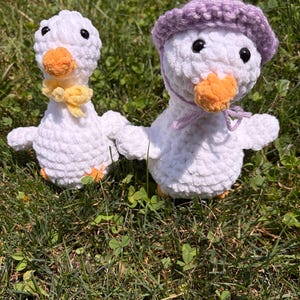 May include: Two white crocheted plush ducks with orange beaks and black eyes. One duck wears a yellow yarn bow around its neck, while the other wears a light purple crocheted bonnet tied with matching strings under its chin.