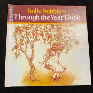 Puede incluir: Un libro de tapa dura titulado Holly Hobbie's Through the Year Book. La portada presenta una sección superior marrón con texto en amarillo y blanco, y una sección inferior blanca con una ilustración de una persona con sombrero en un columpio de árbol.