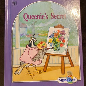 Peut inclure: Un livre pour enfants à couverture rigide intitulé Queenie's Secret avec une bordure violette. L'illustration de couverture montre un oiseau portant un manteau rose, tenant un pinceau et une palette, peignant un vase de fleurs sur un chevalet. Les logos Grolier et AlphaPets sont visibles.