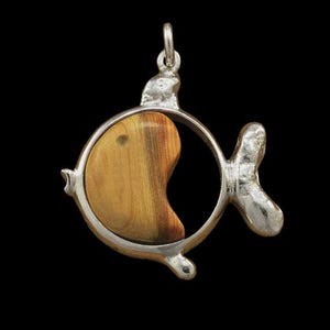 May include: A silver-toned fish pendant with a wooden inlay. The fish-shaped pendant has a circular frame with a wooden insert in the shape of a kidney bean. The pendant has a loop at the top for a chain.