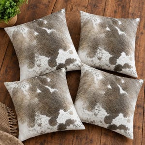 May include: A set of four square throw pillows featuring a brown and white cowhide print pattern. Each pillow has a soft, textured appearance with irregular patches of dark brown and white, creating a natural hide aesthetic on all sides.