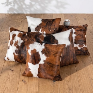 May include: A collection of four rectangular and square throw pillows featuring a cowhide pattern with patches of white, brown, and black fur. The pillows are arranged in a cluster, showcasing the natural texture and varied color distribution of the hide.