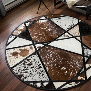 Puede incluir: Una alfombra circular construida con paneles de piel de vaca en tonos marrón, blanco y negro. La alfombra presenta un patrón geométrico de tiras de cuero negro que se cruzan, dividiendo las secciones de piel en varias formas triangulares.
