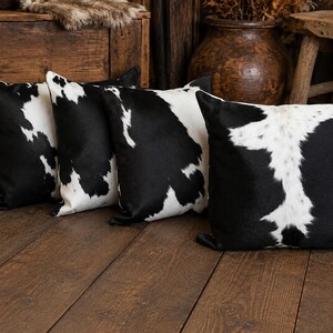 May include: A set of four square throw pillows featuring a black and white cowhide pattern. Each pillow displays a unique arrangement of irregular black patches against a white background, creating a high-contrast, spotted appearance across the fabric.