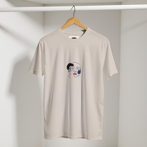 Könnte beinhalten: Ein hellbeige T-Shirt mit einem minimalistischen Gesichtsdesign. Das Design zeigt schwarze, blaue und rote Linienkunst, darunter ein halbes Gesicht mit einem schwarzen Abschnitt, einem blauen Kreis und roten Lippen. Das Shirt hängt an einem Holzbügel.