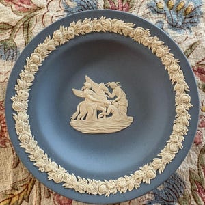 May include: A blue decorative plate with a cream-colored floral border and a central relief scene. The relief depicts figures and a winged horse. The plate's design is classic and elegant, suitable for display.