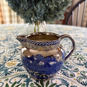 May include: A small ceramic creamer pitcher featuring a metallic gold finish on the upper portion and a vibrant blue and white transferware landscape pattern on the lower body, complete with a decorative blue wave-patterned rim border.