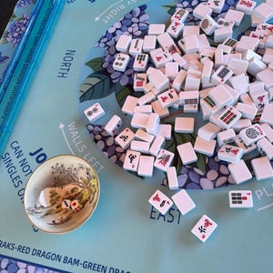 May include: A Mahjong game set featuring white tiles with red, green, and black symbols scattered on a light blue mat with hydrangea patterns. A small ceramic bowl contains two dice, and a blue plastic rack sits on the left side of the board.