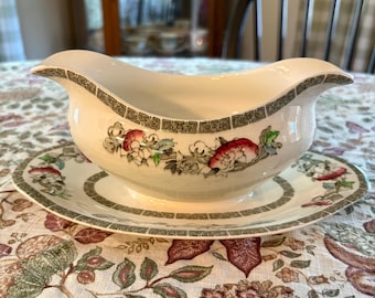 May include: A cream-colored ceramic gravy boat and matching oval underplate. The set features a decorative green geometric border pattern along the rims and a floral motif with red and green accents painted on the sides of the boat and plate.