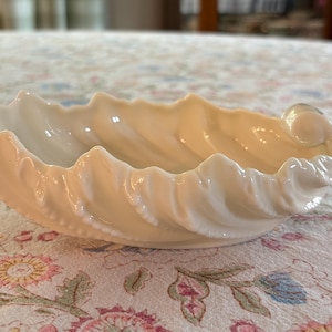 May include: A cream-colored ceramic dish shaped like a seashell with a scalloped, ribbed edge and a curled end. The bowl features a glossy finish and a curved, organic form that mimics the natural texture and structure of a marine shell.