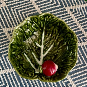 May include: A ceramic bowl shaped like a large, textured green cabbage leaf with a glossy finish. The bowl features a prominent, three-dimensional red radish resting near the bottom center, with white veins embossed across the dark green leaf surface.