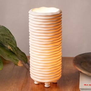 May include: A cream-colored, cylindrical table lamp with a textured, ridged design. The lamp has a warm glow emanating from the top and rests on three small, rounded feet. The lamp is on a wooden surface.