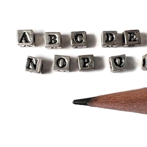 Könnte beinhalten: Silberne Alphabet-Buchstabenperlen mit schwarzer Beschriftung, in zwei Reihen angeordnet, von A bis V, mit einem grünen und braunen Bleistift darunter.