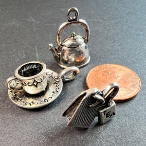 May include: Miniature silver-toned charms including a teapot, teacup and saucer, and a shopping bag. A copper penny is included for size comparison. The charms are detailed and appear to be made of metal.