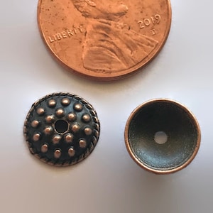 May include: Two antique copper-toned metal beads. One bead is round and has a domed top. The other bead is flat and has a raised pattern of small circles.