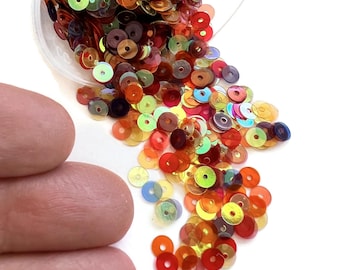 May include: A large assortment of iridescent round sequins in various shades of red, orange, yellow, and green. The sequins are scattered on a white surface.