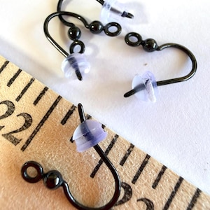 May include: Three black metal fishhook earring wires with small spherical beads and clear silicone backings. A wooden ruler is visible in the frame, showing the earring components measure approximately 0.75 inches in total length.