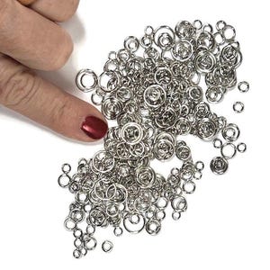 Op de afbeelding: Een verzameling kleine, ronde, zilverkleurige metalen buigringen met variërende diameters. De ringen liggen los verspreid en hebben een gepolijste, reflecterende afwerking. Deze metalen onderdelen zijn bedoeld voor het maken van sieraden.