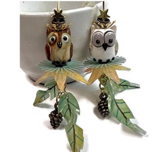 May include: A pair of whimsical owl earrings. Each earring features a detailed owl charm with a star-shaped crown, a frilly collar, and dangling leaf and pinecone accents. The text "ONLINE CLASS" is at the top.