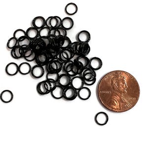 May include: A collection of small, black metal jump rings scattered on a white surface, next to a US penny for size comparison. The rings are circular and appear to be made of a shiny metal. The penny shows the profile of Abraham Lincoln and the year 2020.