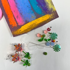 May include: A pair of handmade earrings featuring a pink and orange flower design with green leaves and beads. The earrings are made with polymer clay and are hanging from silver hooks.