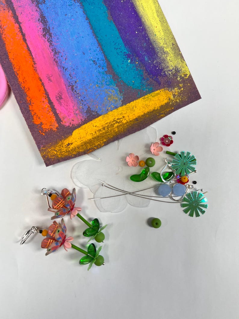 May include: A pair of handmade earrings featuring a pink and orange flower design with green leaves and beads. The earrings are made with polymer clay and are hanging from silver hooks.