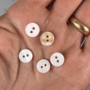 May include: Five white and cream colored buttons with two holes each. The buttons are made of mother of pearl and have a slightly iridescent sheen.