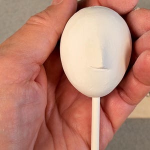 Può includere: Una scultura ovale bianco opaco caratterizzata da un naso leggermente in rilievo e una linea orizzontale per la bocca. L'oggetto è fissato a un sottile bastoncino cilindrico bianco. Sullo sfondo è visibile un righello con scala in centimetri.