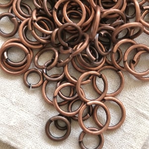 Op de afbeelding: Een verzameling van talrijke cirkelvormige buigringen van koperkleurig metaal. Elke individuele ring heeft een kleine, zichtbare opening langs de omtrek, waardoor ze gemakkelijk kunnen worden verbonden en gemonteerd.