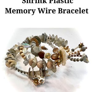May include: A memory wire bracelet with a variety of beads and decorative elements. The bracelet features a mix of neutral colours, including beige, brown, and grey, with floral and shell-like accents. The text "FREE TUTORIAL Shrink Plastic Memory Wire Bracelet" is at the top.