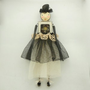 May include: A black and white doll with a black hat and a white dress. The doll is holding a small white frame with a yellow flower inside. The doll is wearing a black tulle skirt and a white lace trim.