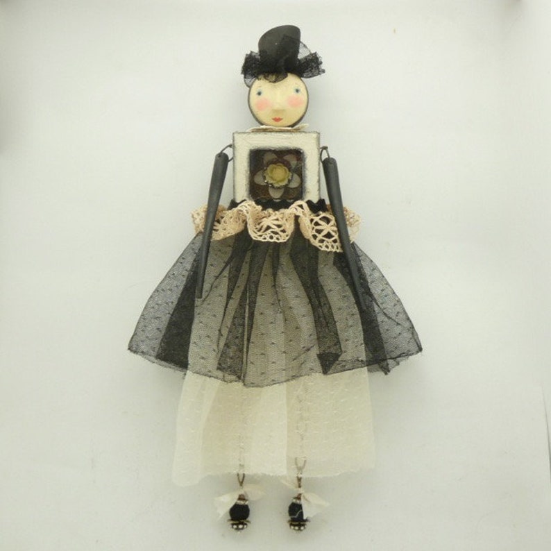 May include: A black and white doll with a black hat and a white dress. The doll is holding a small white frame with a yellow flower inside. The doll is wearing a black tulle skirt and a white lace trim.