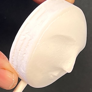 Puede incluir: Un objeto redondo, blanco, con acabado mate y un perfil esculpido de un rostro en su superficie, unido a un palo cil&iacute;ndrico, fino y blanco. El objeto tiene una apariencia texturizada y porosa con un relieve que representa una nariz y una barbilla.