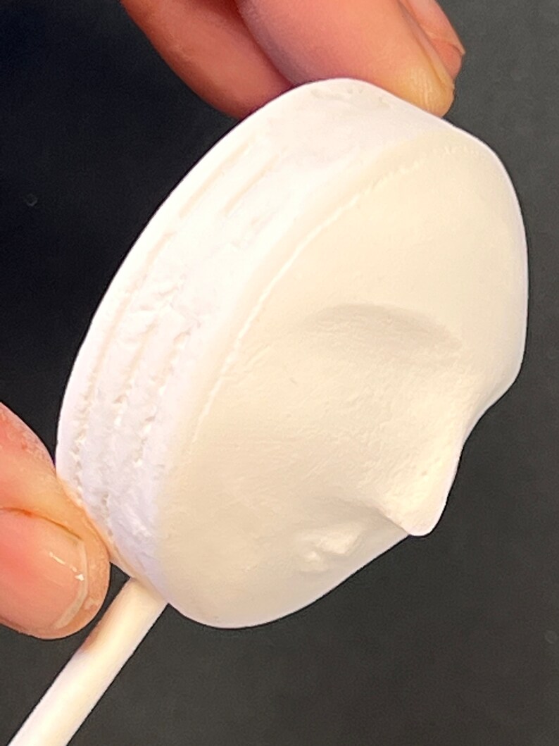 Puede incluir: Un objeto redondo, blanco, con acabado mate y un perfil esculpido de un rostro en su superficie, unido a un palo cil&iacute;ndrico, fino y blanco. El objeto tiene una apariencia texturizada y porosa con un relieve que representa una nariz y una barbilla.
