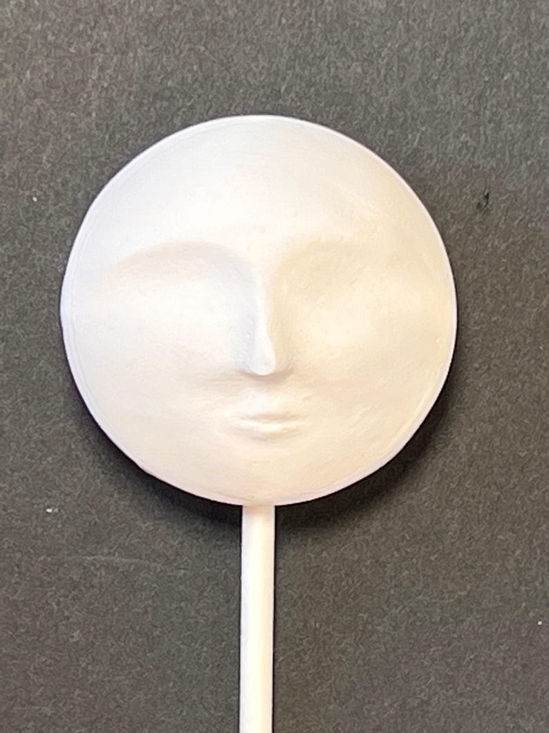 A plain white round plaster doll head with molded facial features attached to a white stick against a dark textured background.
