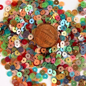 May include: A collection of small, round, multi-colored sequins in shades of red, orange, yellow, green, blue, and pink, scattered around a single United States one-cent copper coin placed in the center for scale.