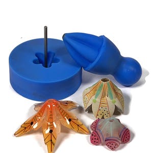 May include: A blue silicone mould with a small metal pin in the centre. The mould is designed to create a flower-shaped object. Three flower-shaped objects are shown, one with orange and yellow accents, one with green and yellow accents, and one with pink and blue accents.