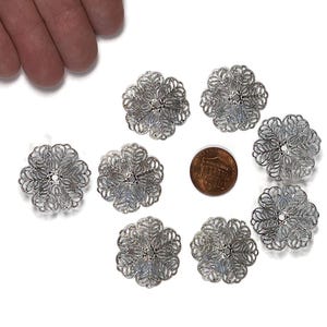 May include: A collection of eight silver-colored, filigree flower-shaped embellishments. Each delicate piece features an intricate openwork design with a central detail. A penny is included for size reference. The items are arranged on a white surface.