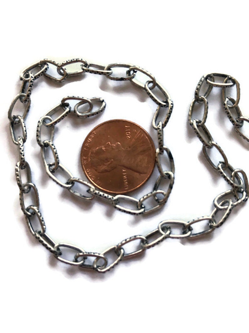 K&ouml;nnte beinhalten: Eine silberne Kette mit strukturierter Oberfl&auml;che. Die Kette ist oval geformt und etwa so gro&szlig; wie ein 1-Cent-St&uuml;ck.