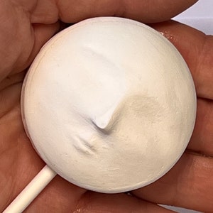 Puede incluir: Un objeto circular de color blanco roto con textura mate que presenta un perfil de rostro humano esculpido en relieve sobre su superficie. La pieza redonda est&aacute; unida a un palo de madera claro, fino y recto, que sale del borde inferior.